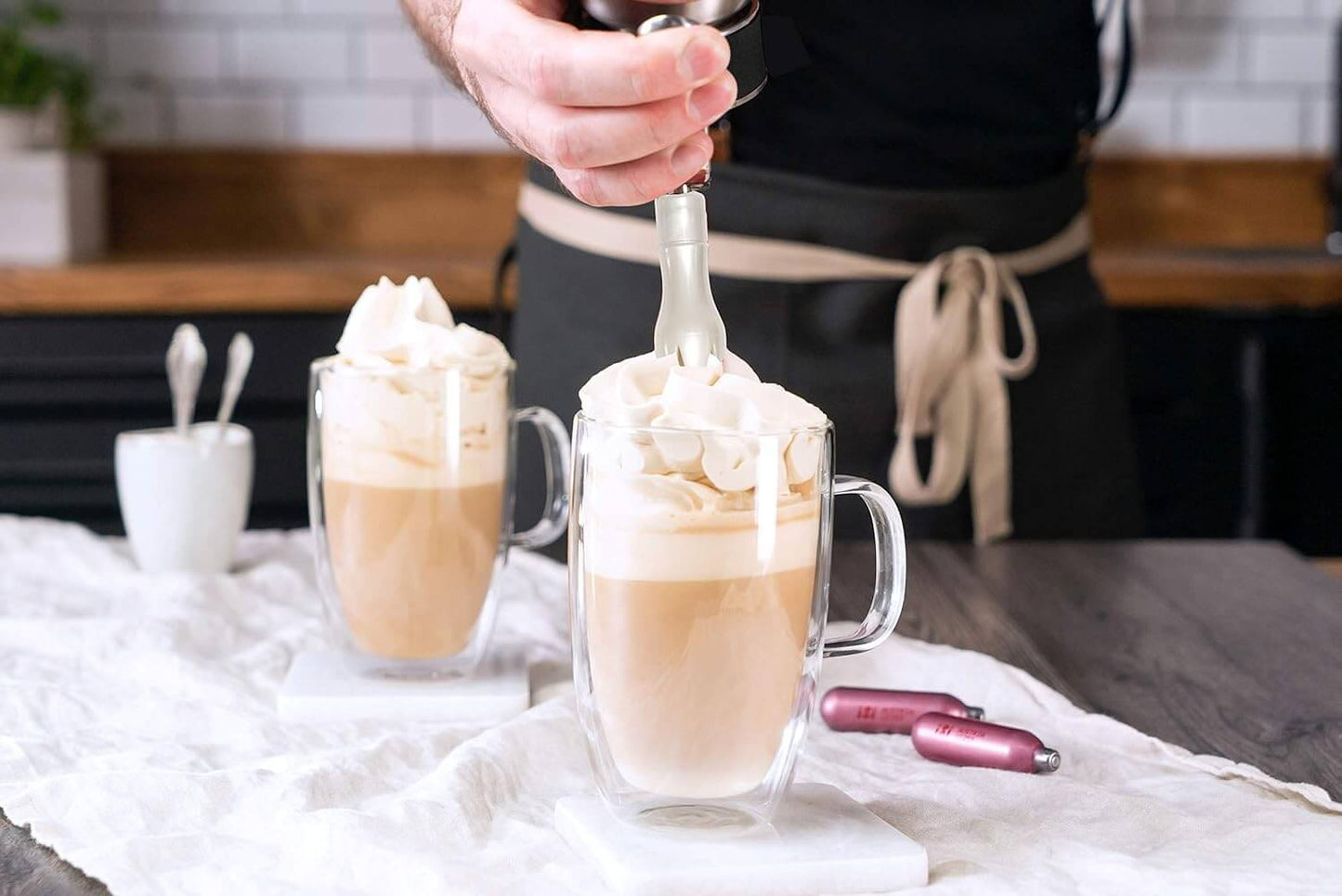 A person in a black apron expertly tops two clear mugs of whipped coffee with extra cream from a stainless steel iSi North America Cream Profi Whip by Vegas Party Favors. The mugs rest on a white cloth on a wooden table, creating an inviting rustic scene.